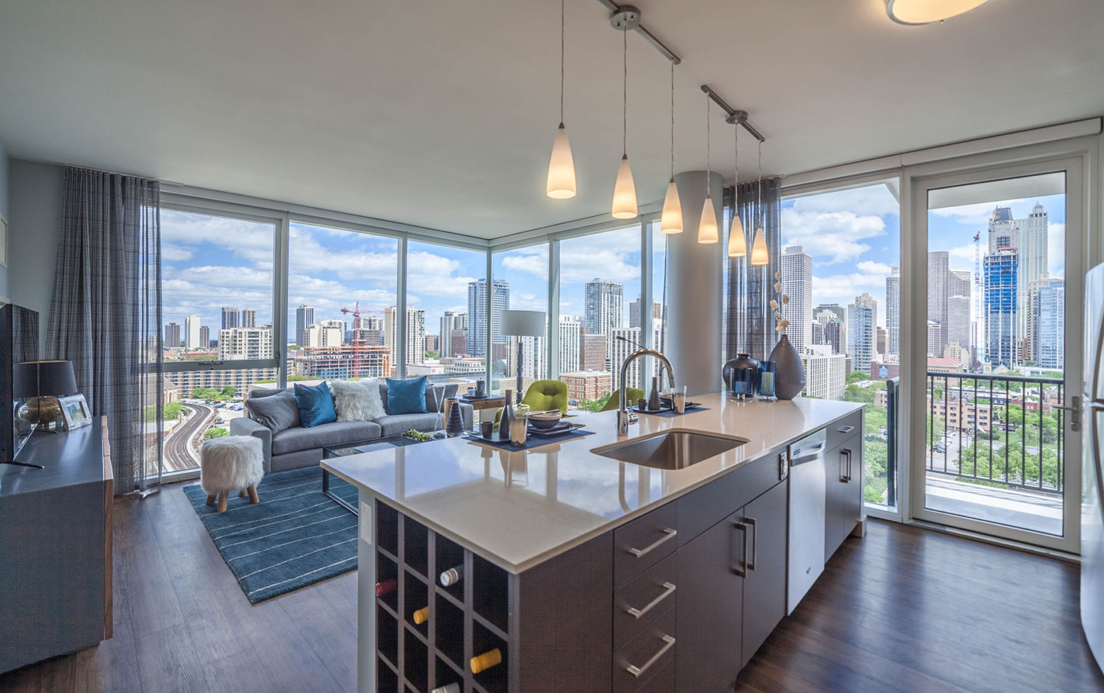 Model kitchen and living room for a two-bedroom apartment with a kitchen island, quartz countertops, plank flooring and floor-to-ceiling windows with a balcony and city view